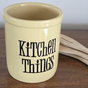 May include: A cylindrical cream-colored ceramic utensil crock with a smooth, glossy finish. The front features the black text Kitchen Things in a stylized, tall font. Two light-colored wooden kitchen utensils are tucked inside the container.