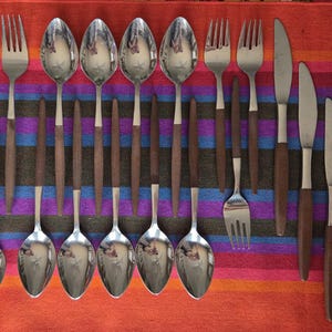 May include: A collection of vintage stainless steel flatware with wooden handles. The set includes knives, forks, and spoons, arranged on a colorful striped textile. The flatware has a mid-century modern aesthetic.