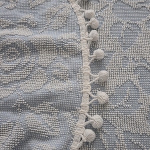 May include: Close-up of a light blue and white patterned textile. The fabric has a textured, dotted appearance with floral designs. A white trim with small pom-poms borders the edge of the textile, adding a decorative element.