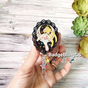 May include: Black and gold anime character badge reel with a moon and stars design. The character is a girl with blonde hair and a pink dress. The reel has a clear plastic strap and a silver moon charm with a pink bead.