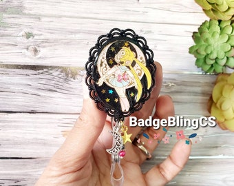 May include: Black and gold anime character badge reel with a moon and stars design. The character is a girl with blonde hair and a pink dress. The reel has a clear plastic strap and a silver moon charm with a pink bead.
