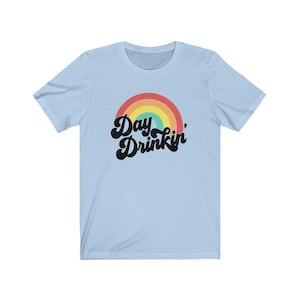 Könnte beinhalten: Hellblaues T-Shirt mit einem Regenbogenbogen und dem Text "Day Drinkin'" in schwarzer Retro-Schrift.