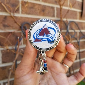 May include: A silver and white retractable badge holder with a round, white, and silver charm featuring the Arizona Coyotes logo. The charm has a red "A" with a blue wave design. The badge holder has a silver chain with a clear plastic holder and a silver and blue hockey stick charm with red, blue, and black beads.