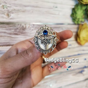 K&ouml;nnte beinhalten: Silberfarbener Anstecker mit einem blauen Strassstein und einem Caduceus-Symbol. Der Anstecker tr&auml;gt die Aufschrift "BSN" in der Mitte.