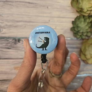 May include: A light blue retractable badge reel with the word "UNSTOPPABLE" and a black dinosaur graphic. The dinosaur is holding a flower. The reel has a clear plastic strap and a black clip.