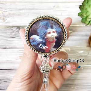May include: A silver and gold badge holder with a round, clear plastic badge holder. The badge holder features a picture of a blue and red creature with the text "Ello!" and a silver teapot charm.