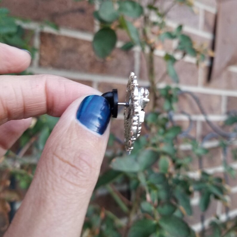 K&ouml;nnte beinhalten: Silberfarbener Ohrring mit einem detaillierten Design. Der Ohrring wird von einem Finger mit blauem Nagellack gehalten.