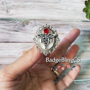 K&ouml;nnte beinhalten: Silberfarbener Anstecker mit einem roten Strassstein, einem Caduceus-Symbol und den Buchstaben "BSN" in einem wei&szlig;en Emaille-Oval. Der Anstecker befindet sich auf einem Finger.