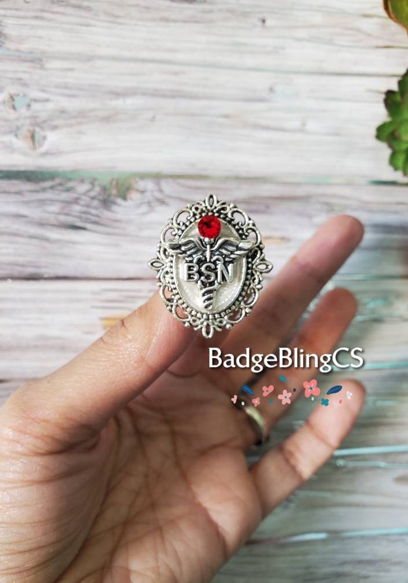 K&ouml;nnte beinhalten: Silberfarbener Anstecker mit einem roten Strassstein, einem Caduceus-Symbol und den Buchstaben "BSN" in einem wei&szlig;en Emaille-Oval. Der Anstecker befindet sich auf einem Finger.