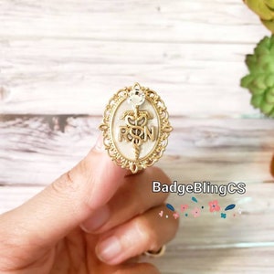 May include: Gold-toned oval-shaped pin with a white background. The pin features a medical caduceus symbol with the letters "RN" in the center. The pin is surrounded by a decorative gold frame.