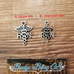 K&ouml;nnte beinhalten: Zwei silberfarbene Charms mit dem medizinischen Symbol einer Schlange, die sich um einen Stab mit Fl&uuml;geln windet, mit den Buchstaben RN auf beiden Seiten. Der Charm links ist gr&ouml;&szlig;er als der Charm rechts. Die Charms befinden sich auf einer Holzoberfl&auml;che mit einer Visitenkarte, auf der "Badge Bling CSS Schmuck f&uuml;r Ihr Arbeitsabzeichen, Broschen und mehr!" steht.