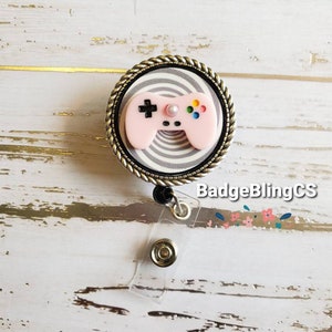 May include: A pink video game controller with black buttons and a white pearl on top, on a black and white striped background, inside a silver and black circular frame with a black rope design. The frame is attached to a clear plastic retractable badge holder with a silver clip.