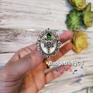 K&ouml;nnte beinhalten: Eine silberfarbene Anstecknadel mit einem gr&uuml;nen Stein in der Mitte. Die Anstecknadel zeigt ein Caduceus-Medizinsymbol mit den Buchstaben "BSN" auf der Vorderseite. Die Anstecknadel ist von einem dekorativen filigranen Rahmen umgeben.