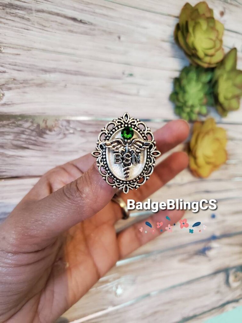 K&ouml;nnte beinhalten: Eine silberfarbene Anstecknadel mit einem gr&uuml;nen Stein in der Mitte. Die Anstecknadel zeigt ein Caduceus-Medizinsymbol mit den Buchstaben "BSN" auf der Vorderseite. Die Anstecknadel ist von einem dekorativen filigranen Rahmen umgeben.