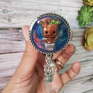 May include: A silver and blue galaxy themed badge reel with a small resin figure of Groot from Guardians of the Galaxy. The reel has a clear plastic badge holder and a silver chain with star and moon charms.