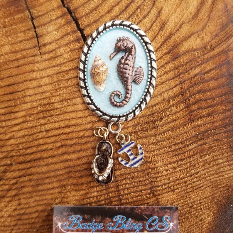 May include: A silver oval charm with a blue background and a brown seahorse and seashell. The charm has a silver rope border and is attached to a silver chain with a gold flip flop and a blue and white anchor charm.