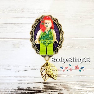 May include: A green and purple Poison Ivy Lego minifigure in a gold ornate frame with a gold leaf charm. The charm is attached to the bottom of the frame. The background is a white wooden surface.