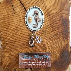 May include: A silver oval charm with a blue background featuring a brown seahorse and a seashell.  The charm is attached to a gold chain with a gold anchor and a pair of gold flip-flop charms.  The text "Badge Bling CS" is on a white card with black text.  The text below reads "Jewelry for your work badge, brooch pins, and more!"