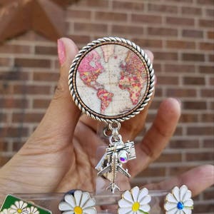 May include: A silver retractable badge holder with a world map design. The map is in shades of pink, brown, and yellow. The holder has a silver airplane charm and a silver camera charm. The holder is attached to a green and white daisy pin.