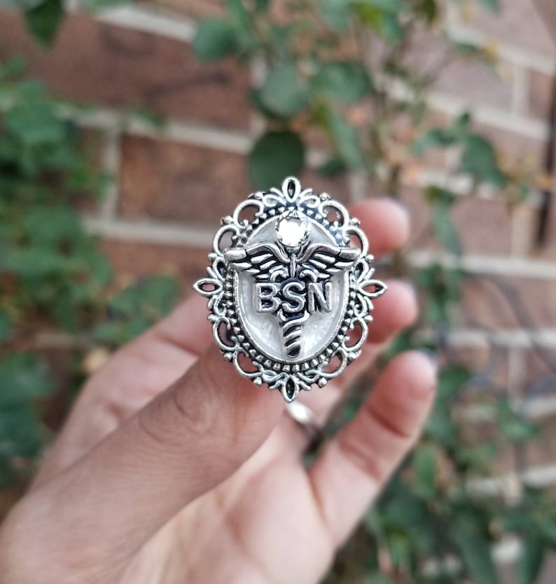 K&ouml;nnte beinhalten: Ein silberner Ring mit wei&szlig;em Hintergrund und einem Caduceus-Symbol mit den Buchstaben BSN. Der Ring ist verziert und hat einen klaren Stein in der Mitte.