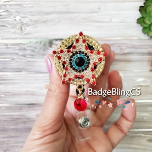 May include: Gold tone star-shaped badge reel with a red, blue, and black eye design. The reel has a red crystal on the end of the retractable cord. The text "BadgeBlingCS" is visible in the image.