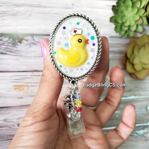 May include: A white oval badge holder with a yellow rubber ducky wearing a nurse's hat. The ducky is surrounded by white and blue glitter. The badge holder has a silver rope border and a clear plastic retractable badge reel with colorful beads.