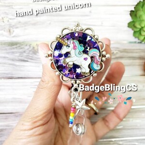 May include: A purple and silver badge reel with a hand-painted unicorn design. The unicorn is white with a pink mane and tail. The background is filled with purple glitter. The reel has a clear plastic badge holder and a rainbow beaded chain with a silver star charm.