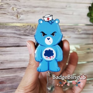 May include: Blue Care Bear with a red cross on a white hat, wearing a nurse's uniform, with a black cloud and hearts on its chest. The Care Bear is attached to a retractable badge reel with a clear plastic strap and a silver spring.