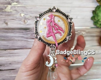 May include: A silver and gold tone badge reel with a pink and white cameo of a girl in a moon and stars design. The reel has a silver and white moon charm with a star on it.