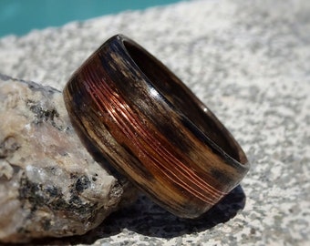 May include: A wooden ring with copper wire inlay. The ring is dark brown with a reddish-brown copper wire inlay. The ring is sitting on a rock.