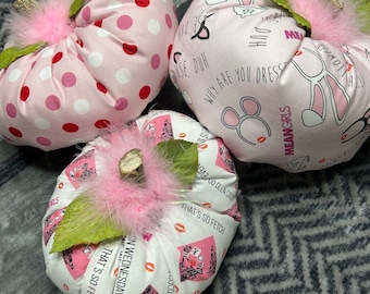 May include: Three pink fabric pumpkins with different patterns. One pumpkin has pink and white polka dots, another has a pink and white pattern with the text "Mean Girls" and the third has a pink and white pattern with the text "On Wednesdays We Wear Pink" and "That's So Fetch".