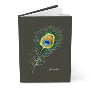 May include: A dark green journal with a watercolor illustration of a peacock feather. The feather has a blue and green eye with a brown center. The name "Jacqueline" is written in cursive at the bottom of the journal.