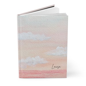 Puede incluir: Un diario de acuarela rosa y azul con un diseño de cielo y nubes. El diario tiene el nombre "Louisa" escrito en la portada.