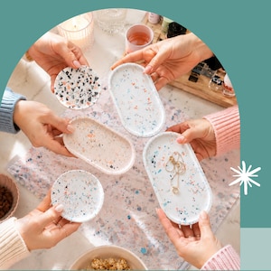 Könnte beinhalten: Eine Sammlung von Terrazzo-Tabletts und Untersetzern in verschiedenen Formen und Farben. Die Tabletts sind oval und rechteckig, während die Untersetzer rund sind. Das Terrazzomuster weist weiße, schwarze, blaue und pfirsichfarbene Sprenkel auf. Ein Tablett enthält eine goldene Halskette.