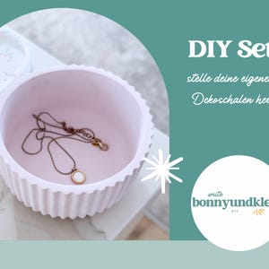 Puede incluir: Un cuenco circular de color rosa claro con bordes acanalados que contiene un collar fino de tono dorado con un colgante circular. El texto "DIY Set" y "stelle deine eigenen Dekoschalen her" es visible, junto al logo "write bonnyundkleid DIY".