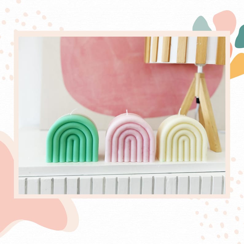 May include: Three arched rainbow-shaped candles arranged in a row. The candle on the left is mint green, the middle candle is light pink, and the candle on the right is pale yellow. Each candle features a single white wick at the top center.
