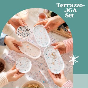 Peut inclure: Un ensemble Terrazzo-JGA compos&eacute; de quatre plateaux d&eacute;coratifs de formes ovales et circulaires. Les plateaux pr&eacute;sentent des bases blanches, beiges et bleu clair avec des motifs mouchet&eacute;s multicolores en noir, orange et bleu. Un plateau contient un collier.
