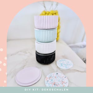 Puede incluir: Una pila de cuatro recipientes cil&iacute;ndricos con texturas acanaladas verticales en rosa pastel, verde menta, blanco y negro. Dos tapas circulares se colocan delante, una de color rosa claro s&oacute;lido y otra con un patr&oacute;n de terrazo moteado multicolor.
