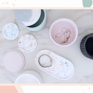 May include: An assortment of decorative trays, coasters, and containers in various colors and designs. Includes a white oval tray with a bracelet, a pink scalloped-edge tray with a necklace, and terrazzo-style coasters.