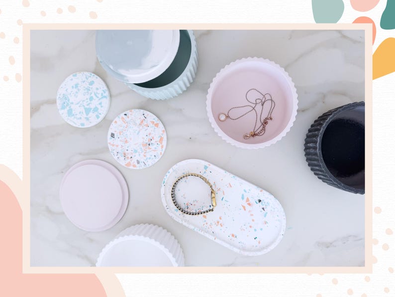 May include: An assortment of decorative trays, coasters, and containers in various colors and designs. Includes a white oval tray with a bracelet, a pink scalloped-edge tray with a necklace, and terrazzo-style coasters.
