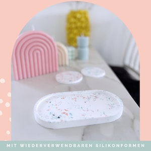 May include: White oval tray with colorful terrazzo-style speckles. A pink rainbow-shaped candle, coasters, and other decorative items are in the background. The text at the bottom reads "MIT WIEDERVERWENDBAREN SILIKONFORMEN".