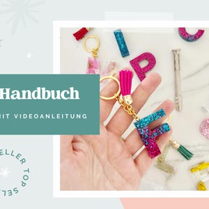May include: A glittery resin keychain in the shape of the letter F, featuring a gradient of blue and magenta, attached to a gold-tone metal ring with a bright pink tassel. Other resin letter charms and a small metal tool are visible nearby.