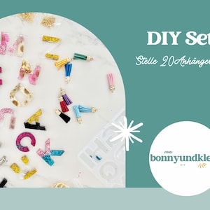 May include: A DIY set for creating 20 charms, featuring various resin alphabet letters with glitter, dried flowers, and gold flakes, alongside colorful fabric tassels with gold caps, a metal hand drill, and a silicone mold on a white surface.