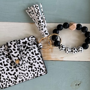 May include: A black and white patterned card holder and keychain wristlet set. The card holder has a scalloped edge and a gold snap closure. The wristlet features black and white spotted beads and a wooden accent.