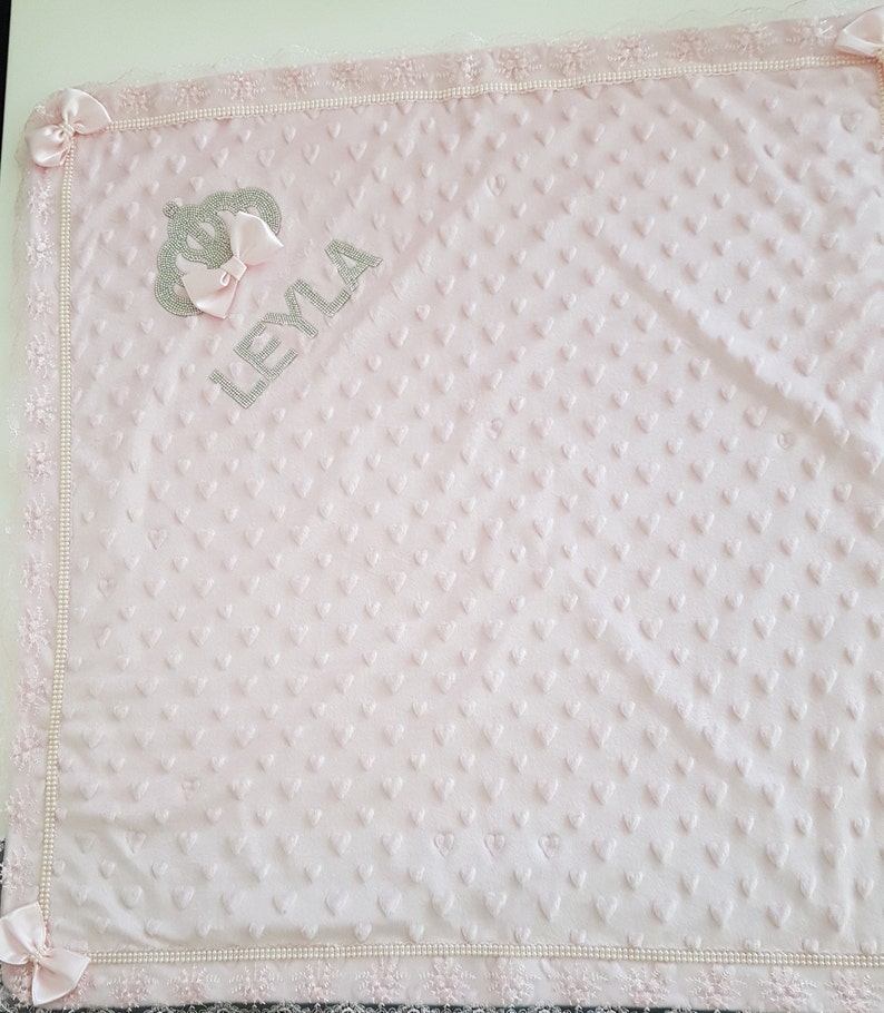 May include: A pink baby blanket with a white lace border and a silver crown and the name "LEYLA" embroidered on it.