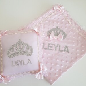 May include: Pink baby blanket and pillow set with a crown and the name "LEYLA" embroidered in silver rhinestones. The blanket has a heart pattern and lace trim.