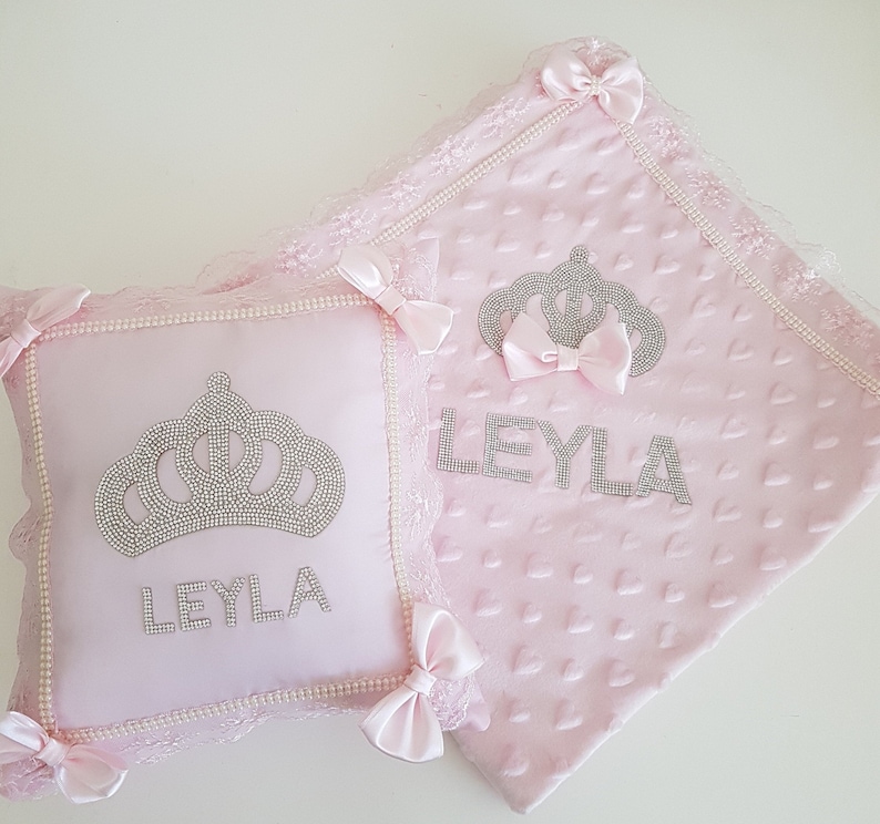 May include: Pink baby blanket and pillow set with a crown and the name "LEYLA" embroidered in silver rhinestones. The blanket has a heart pattern and lace trim.