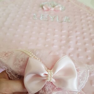 May include: A pink baby blanket with a white lace trim and a pink satin bow. The blanket has a quilted texture and the name "LEYLA" is embroidered on it.