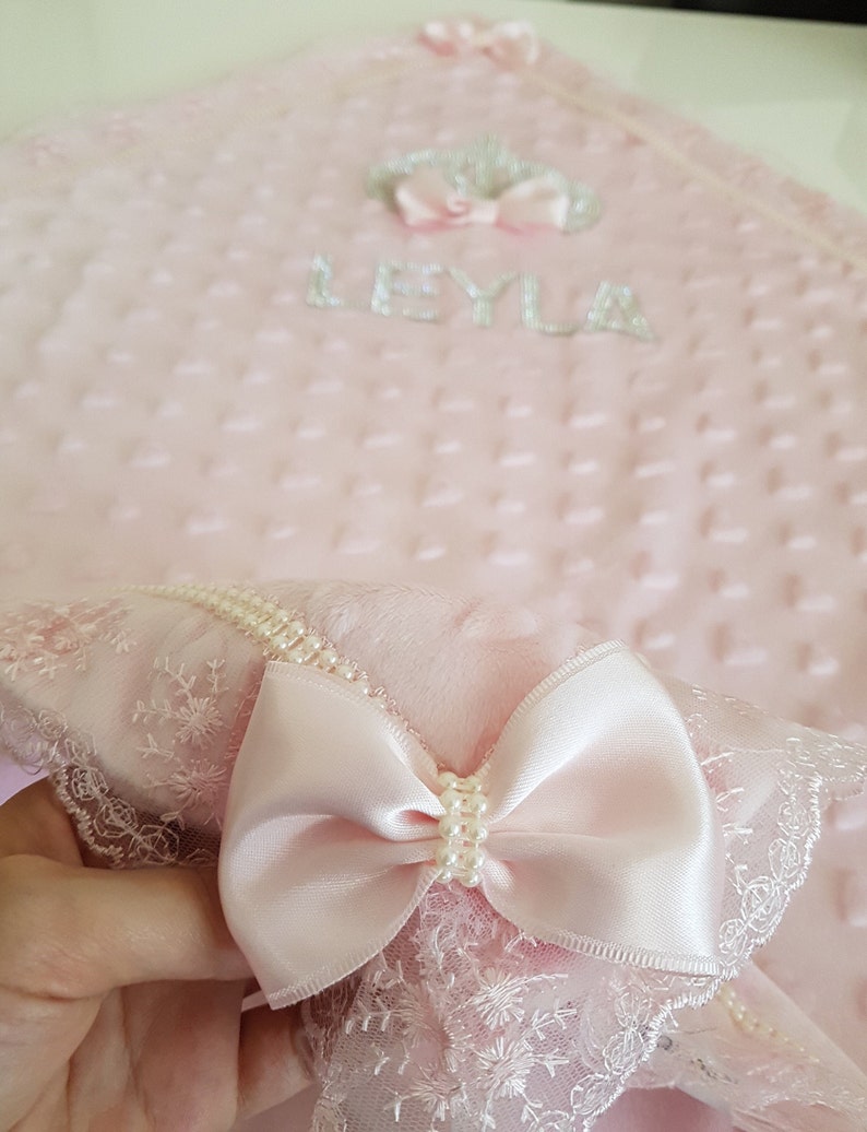 May include: A pink baby blanket with a white lace trim and a pink satin bow. The blanket has a quilted texture and the name "LEYLA" is embroidered on it.