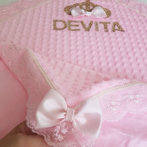 May include: A pink baby blanket with a white lace trim and a gold crown with the name "DEVITA" embroidered on it.